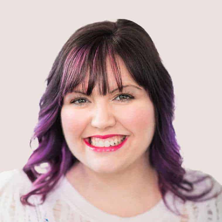 Smiling woman with purple and black hair, bright makeup, and white top.